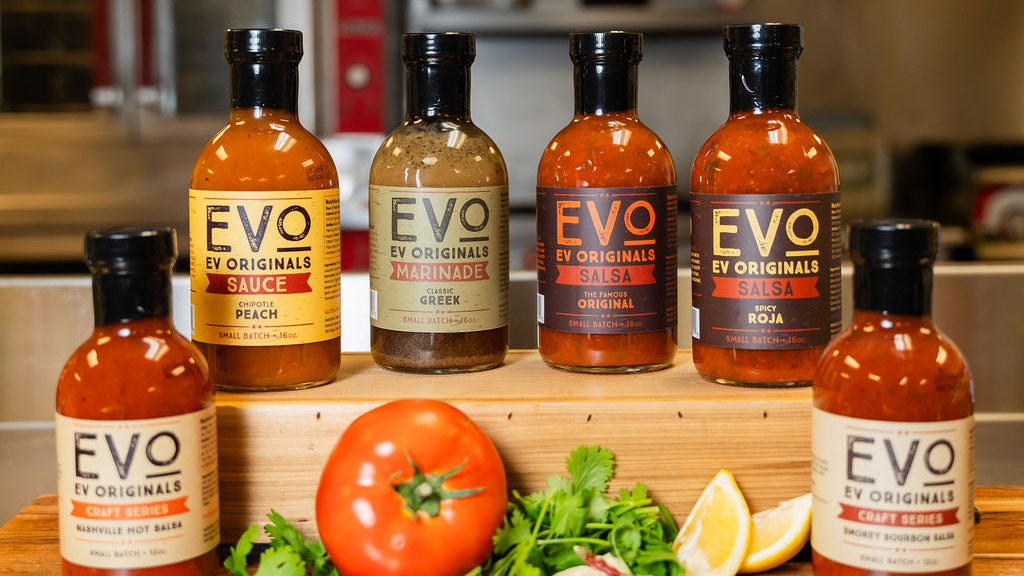 Collection of EVO Originals sauce bottles on a wooden block with fresh vegetables in a kitchen setting.