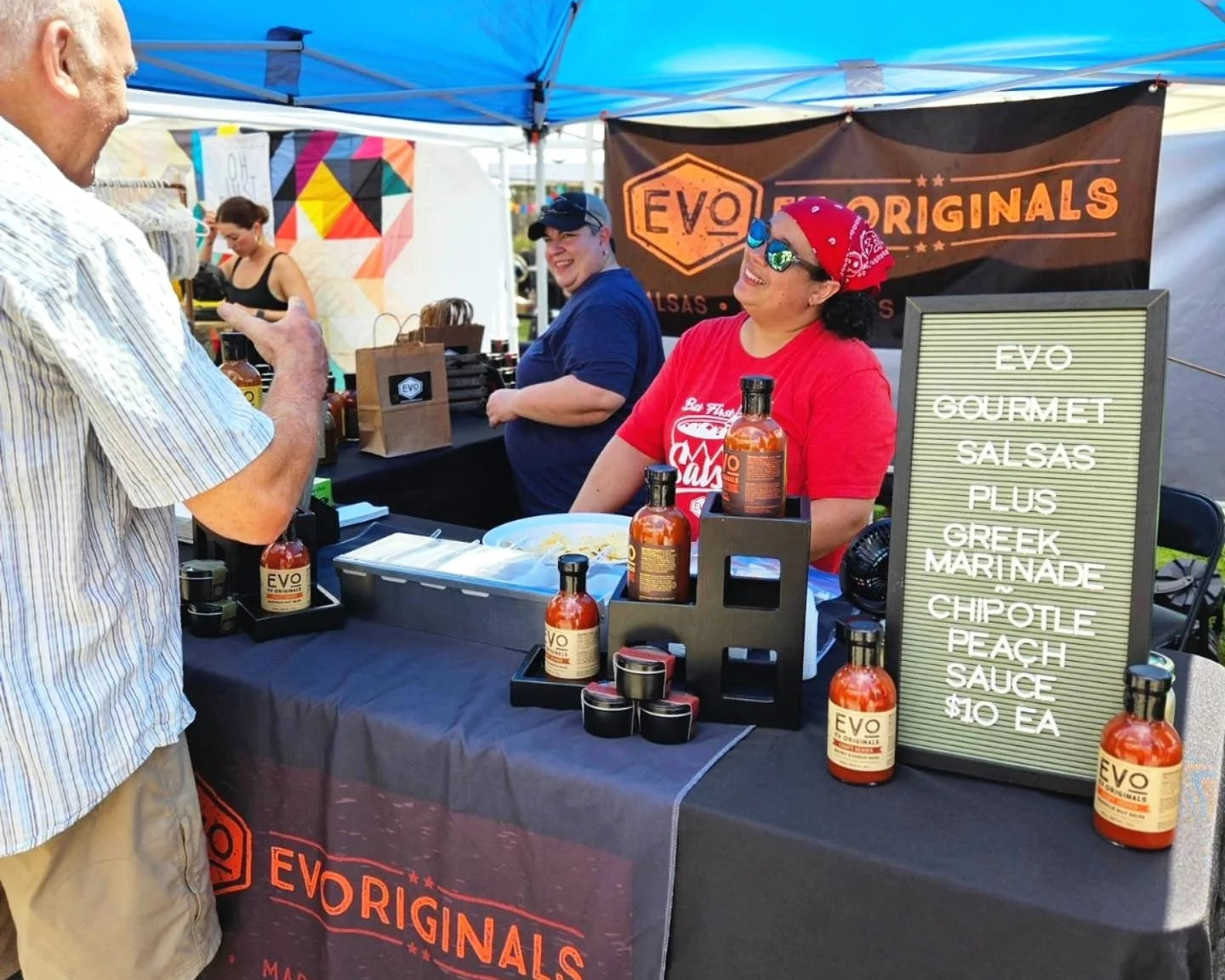 Evo Originals food stand with various sauces and a sign listing products at an outdoor event.